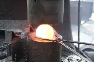 Glowing steel ball being forged under a hydraulic hammer during the industrial production process.