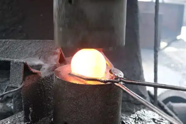 Glowing steel ball being forged under a hydraulic hammer during the industrial production process.
